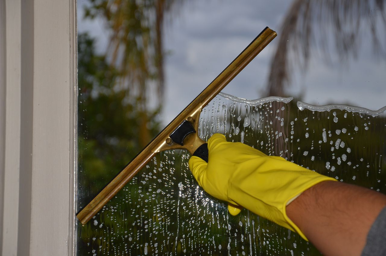 hero-img Close-up of hand cleaning window with squeegee outdoors using yellow gloves.